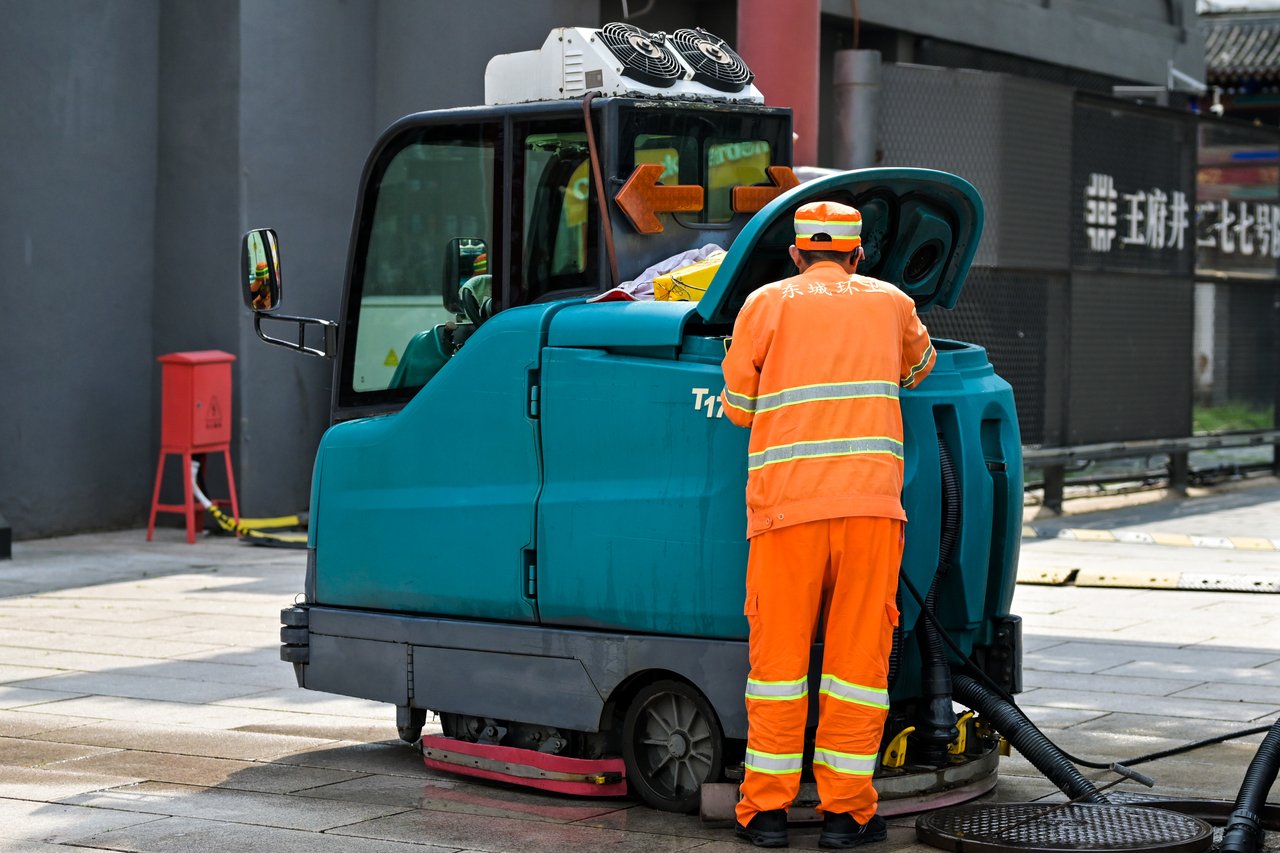 A street cleaner in uniform operates a cleaning machine on an urban sidewalk.